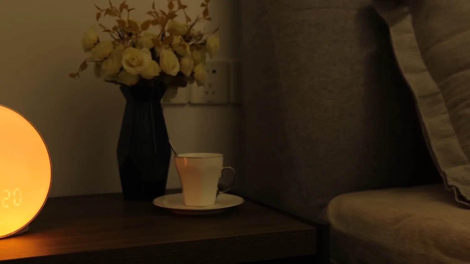 A JALL sunrise alarm clock glowing amber on a nightstand beside a vase of flowers